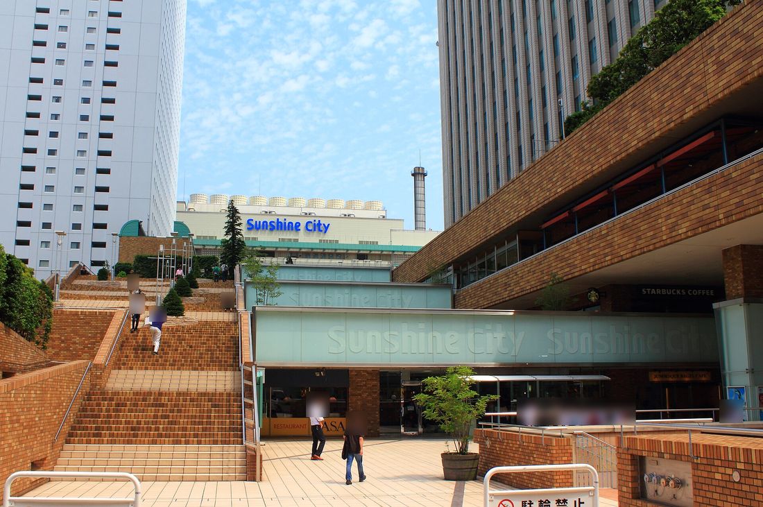 陽光城主建築的入口廣場：左邊是往上的紅磚大階梯，正中間淡綠色玻璃外牆上刻著 Sunshine City 字樣，後方建築頂上有藍色 Sunshine City 招牌，右邊是紅磚外牆的高樓與 STARBUCKS COFFEE 招牌，晴天