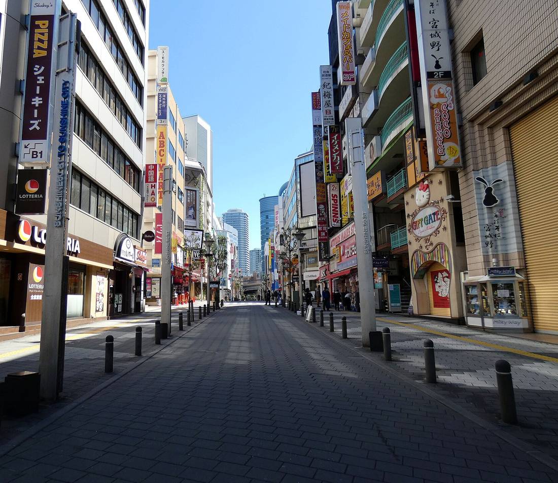 晴天、幾乎沒有行人的サンシャイン60通り：深灰色石磚步行街兩側是密集的招牌，左邊有 LOTTERIA 與 PIZZA シェーキーズ，右邊有 KFC 與 GIFT GATE，街道盡頭遠處是一棟藍色玻璃圓柱形高樓