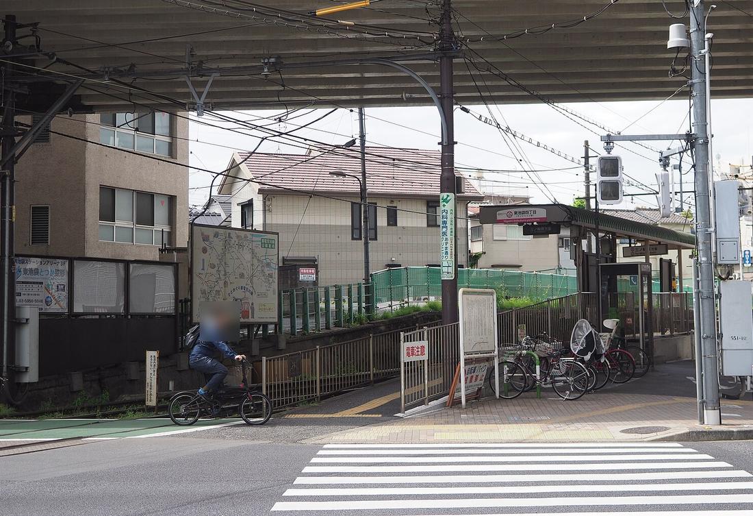 高架快速道路底下的都電荒川線東池袋四丁目電停：右邊是有綠色屋頂的月台與「東池袋四丁目」站名牌，旁邊停著幾台腳踏車，前景是斑馬線，左邊一位騎腳踏車的人