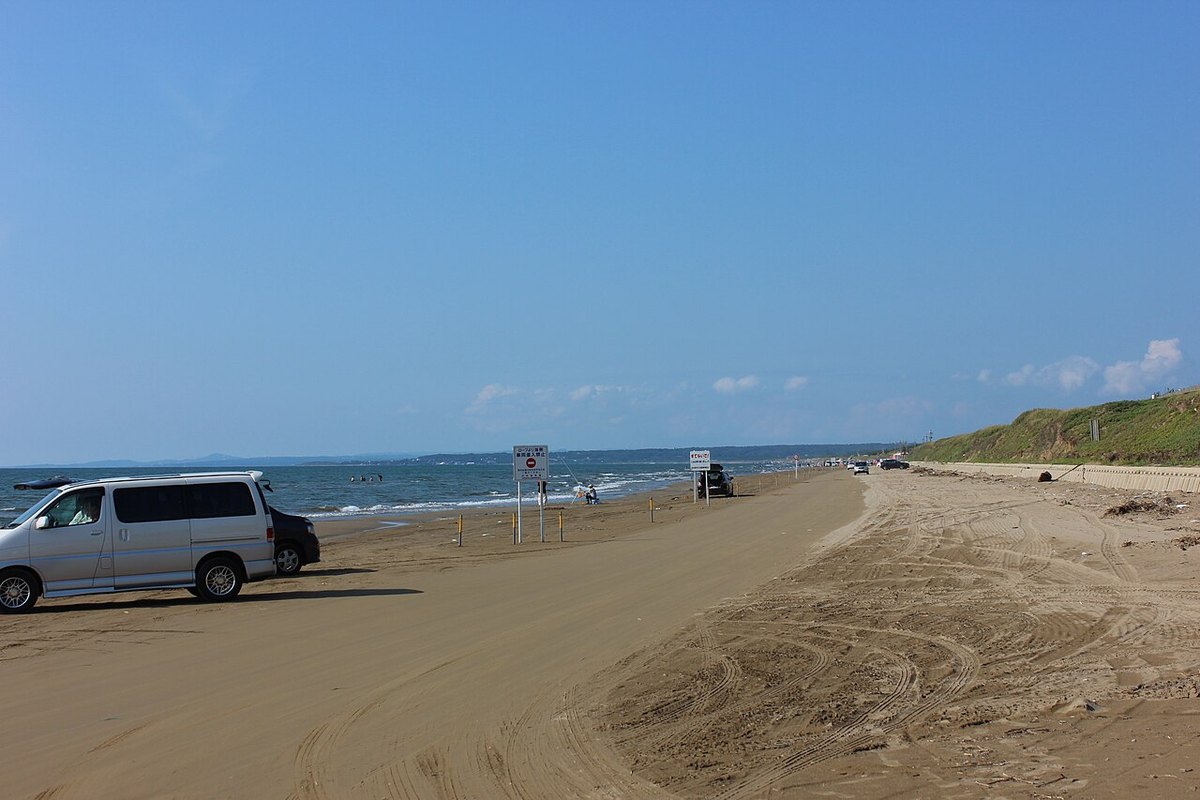Chirihama Nagisa Driveway: a broad stretch of wave-packed sand used as a road, covered in tyre tracks, a van parked at the left, a few more cars and anglers further along the beach, grassy dunes to the right and a deep blue sky
