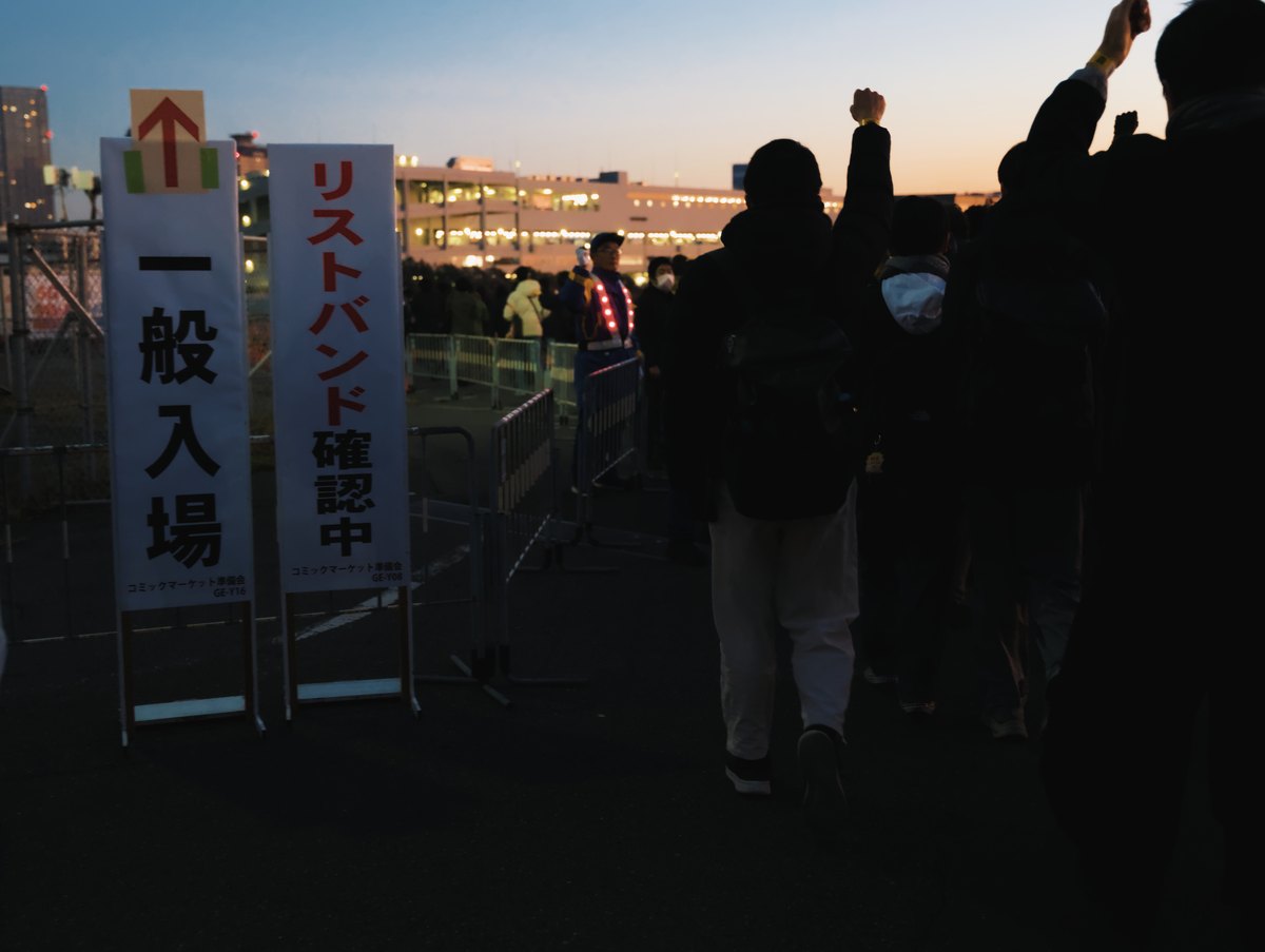 黎明時分的排隊入口:鐵欄杆旁立著兩塊白色看板,左邊黑字「一般入場」上方貼紅色箭頭,右邊紅黑字「リストバンド確認中」,穿紅色發光背心的工作人員站在欄杆通道口,排隊的人背對鏡頭往裡走,有人舉起戴著手環的手