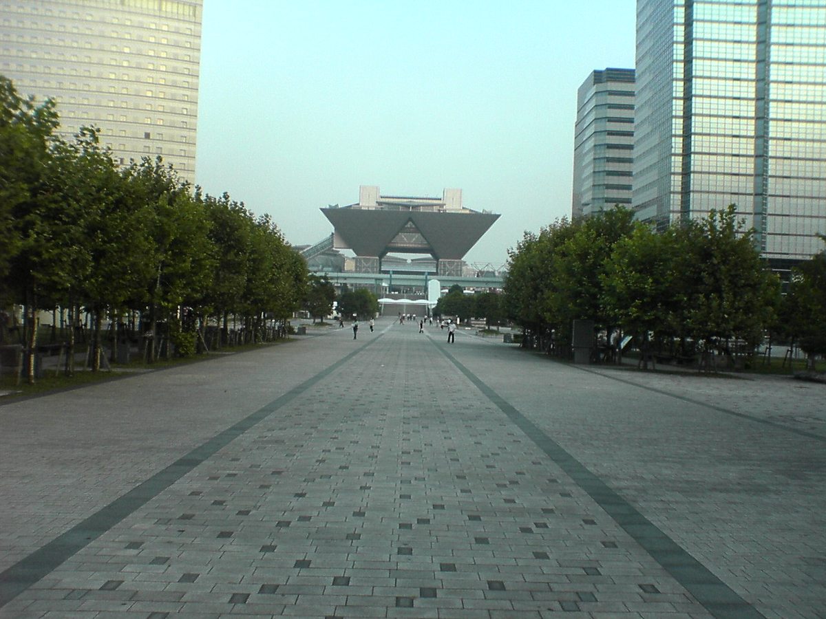 臨海線國際展示場站前往東京 Big Sight 的寬闊石板步道,兩旁是行道樹與玻璃帷幕高樓,步道盡頭是逆三角形的會議棟,路上只有零星行人