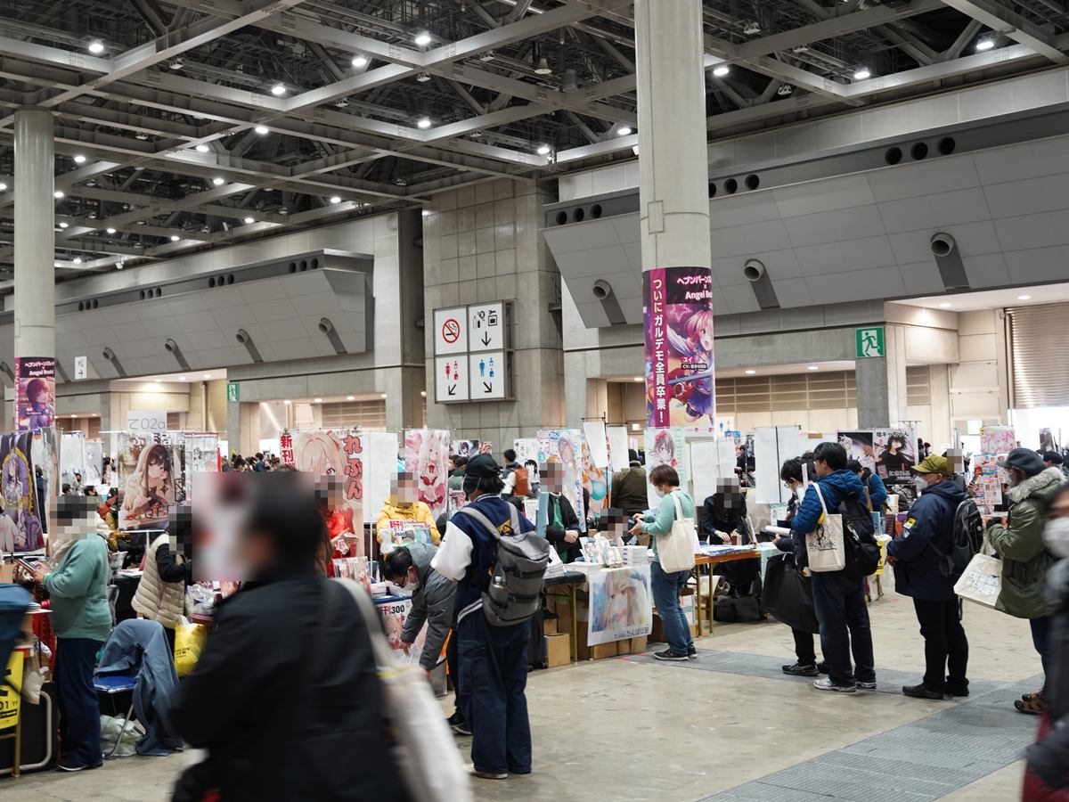 冬季 Comiket 西展示棟的社團攤位區:挑高鋼骨天花板下,一張張長桌排成攤位,桌後立著動漫插畫海報與直幅布條,買家背著後背包、提著帆布袋在攤位前排隊,柱子上掛著「ついにガルデモ全員卒業」的宣傳布條