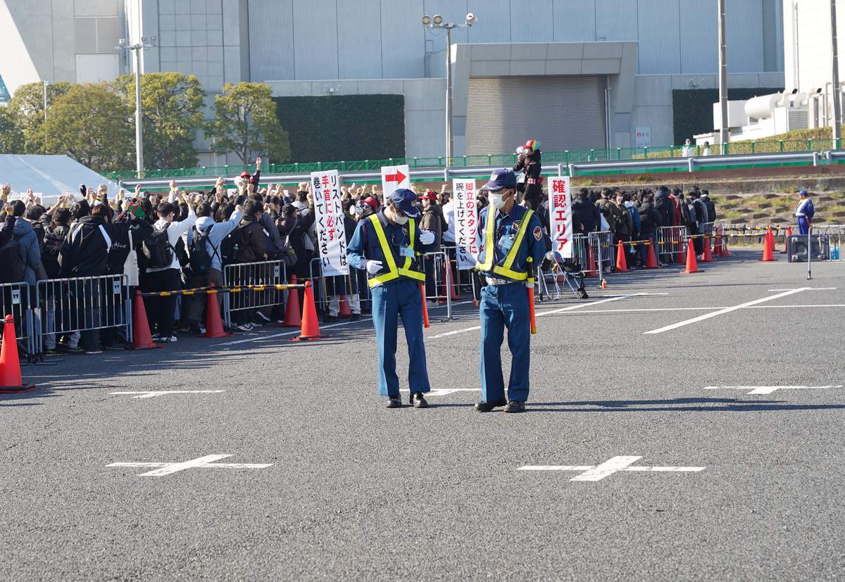 戶外手環確認區:兩名戴口罩、穿反光背心的保全站在空地上,後方欄杆內的入場人群一起舉起手臂,人群前立著看板「リストバンドは手首に必ず巻いてください」「脚立のスタッフに腕を上げてください」和紅字「確認エリア」,一名工作人員站在梯子上拿擴音器