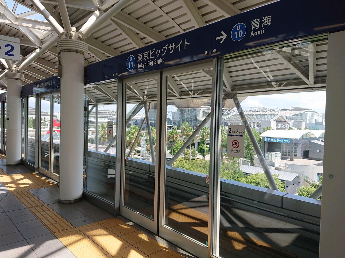 百合海鷗號東京ビッグサイト站的月台:月台門上方的深藍色站名牌寫著 U11「東京ビッグサイト Tokyo Big Sight」,右邊箭頭指向下一站 U10「青海 Aomi」,玻璃月台門外是棕櫚樹與展館建築