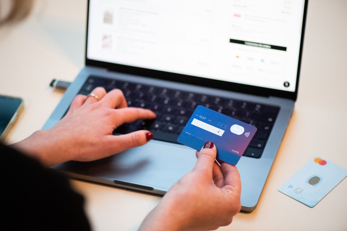 One hand holding a physical bank card while the other types at a laptop showing an online checkout page, a second card with a visible IC chip and contactless symbol lying on the desk