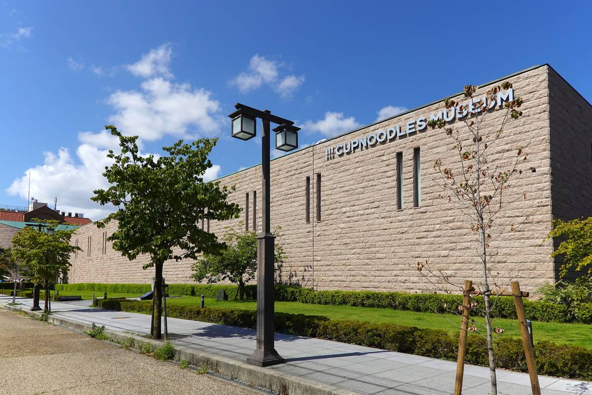 大阪池田杯麵博物館外觀:一長排淺米色石材外牆、只有幾道細長窗,牆上方寫著 CUPNOODLES MUSEUM,前面是草坪、低矮樹籬與人行道路燈