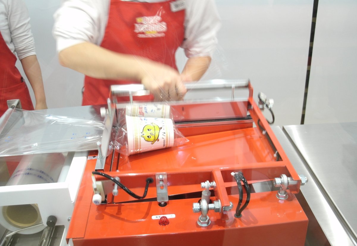 池田館我的杯麵工廠的封膜機:穿紅色 CUP NOODLES 圍裙的工作人員把一個畫了黃色小雞的杯麵放進透明塑膠膜,紅色機台上方是封膜用的金屬桿