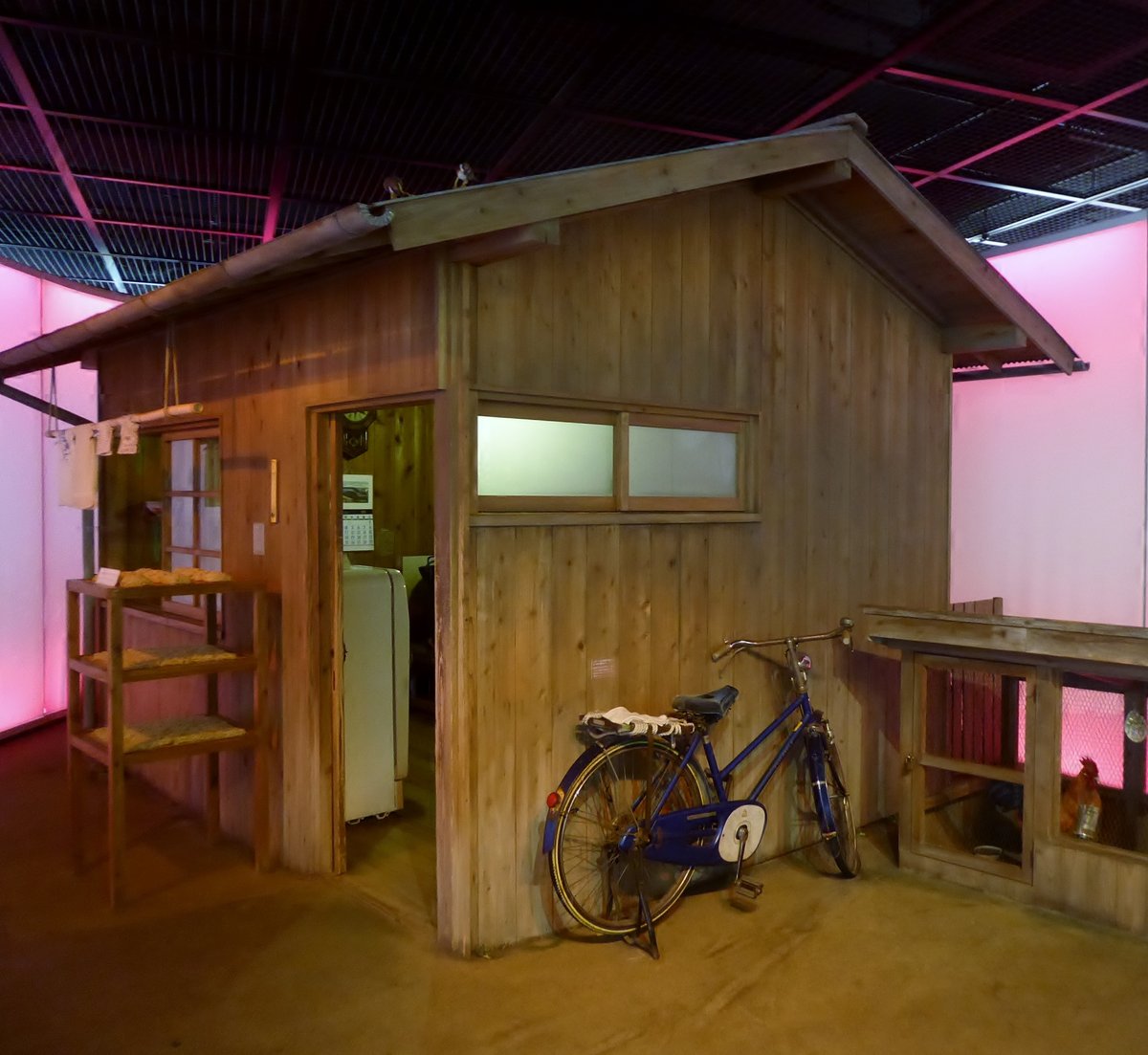 池田館館內重現的安藤百福研究小屋:木板搭的小平房,門口看得到裡面的舊冰箱與月曆,左邊木架上擺著麵塊,牆邊靠著一台藍色舊腳踏車,右邊是一座木造雞舍