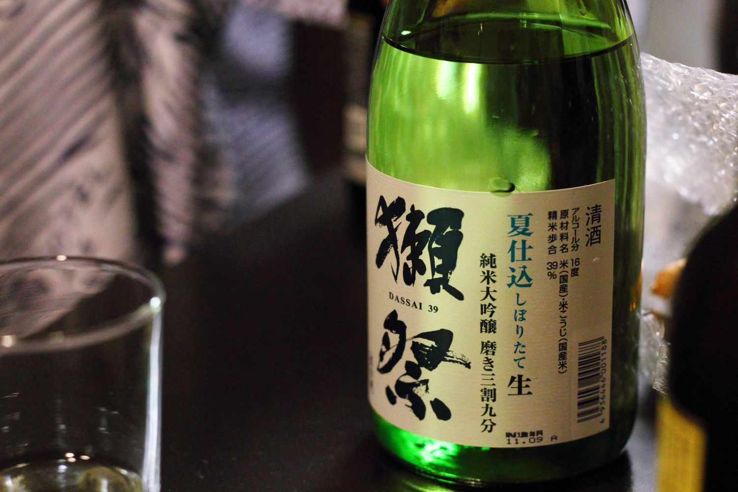 一支綠色玻璃的獺祭酒瓶擺在深色桌面上,瓶身貼著米白色直式標籤,右起以毛筆體寫著大大的「獺祭」二字,下方小字印著 DASSAI 39;標籤中段由右至左直排著「夏仕込 しぼりたて 生」與「純米大吟醸 磨き三割九分」,其中「生」字單獨一個字、字級明顯放大,再往右是清酒、原材料名、精米歩合 39%、アルコール分 16 度等成分欄與條碼。畫面左前方有一只空的玻璃杯,右側可以看到一疊氣泡布
