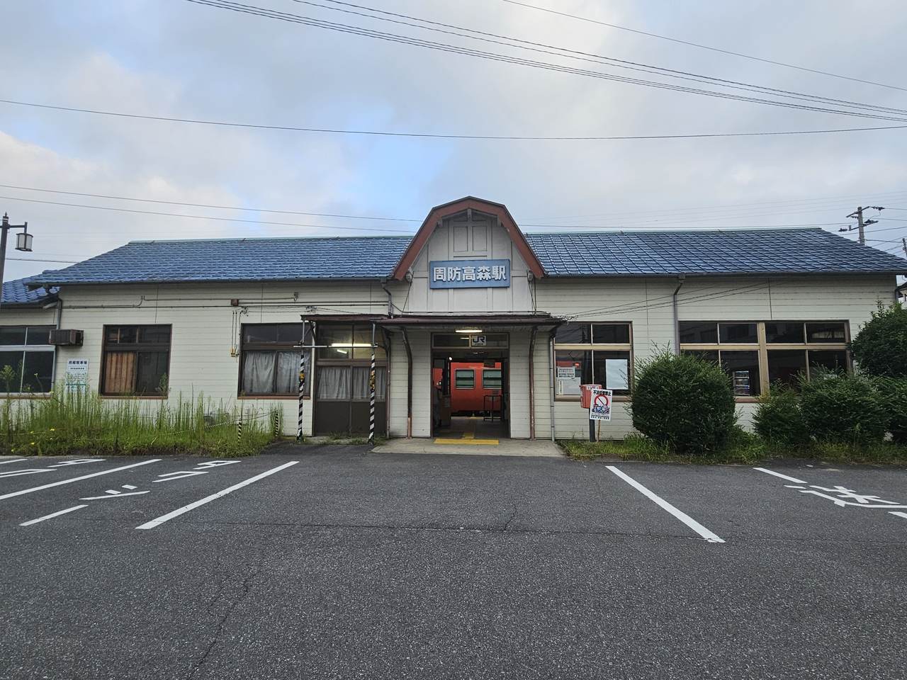 周防高森駅站體正面:一棟米白色木造平房,藍色瓦片屋頂,正中央的三角形山牆上掛著藍底白字的「周防高森駅」站名牌,入口門楣上是白色的 JR 標誌;穿過敞開的出入口可以看到月台上停著一輛朱紅色柴油列車。屋子兩側的窗前長著及腰的雜草,右側立著一塊紅白色的告示牌。建物前方是一片畫著白色停車格線的柏油空地,格子裡一輛車都沒有,也沒有任何排班車輛