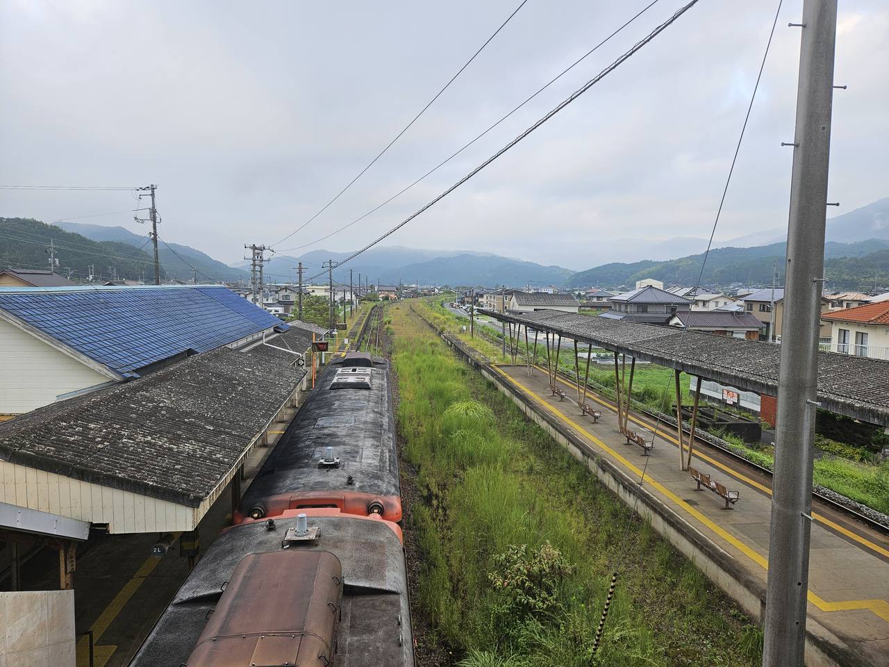 從跨線橋上俯瞰周防高森駅:畫面左下是兩節朱紅色柴油列車停在藍瓦站房旁的月台邊,右側是另一座老舊木柱雨棚的月台,上面排著幾張長椅、空無一人;兩座月台之間的軌道與空地長滿及膝的綠色雜草。再往遠處是低矮民宅組成的小鎮,一路延伸到被雲霧壓著的連綿山稜,天空是灰白色的陰天