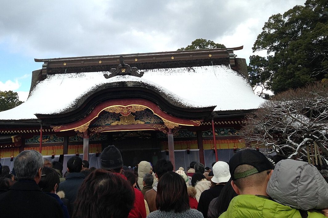 2015 年元旦下午的太宰府天滿宮本殿前：屋頂積著雪的本殿，正面是朱紅與金色裝飾的弧形屋簷，前方整片是穿著羽絨外套、戴毛帽排隊的人群背影，右邊是枝條積雪的梅樹