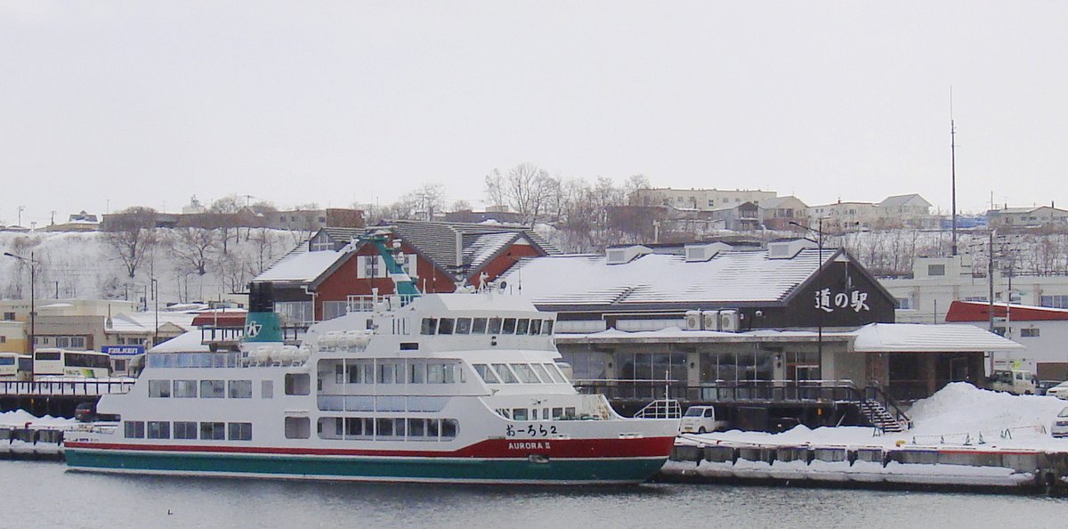 Michi-no-Eki Ryuhyo-kaido Abashiri seen across the Abashiri River: a snow-roofed building with a white roadside-station sign, and a white sightseeing icebreaker with a red-and-green waterline moored at the quay in front, its hull lettered Aurora II