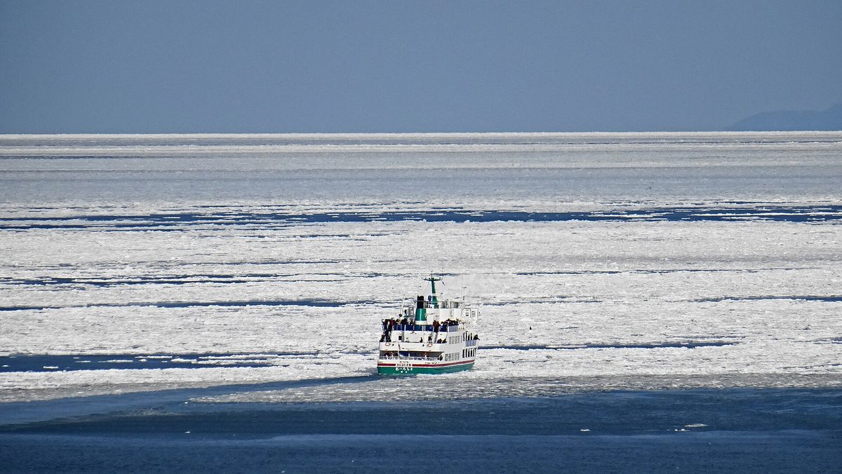 The sea off Abashiri seen from high ground: a white-and-green sightseeing icebreaker crossing dark blue open water into a white belt of drift ice that runs to the horizon