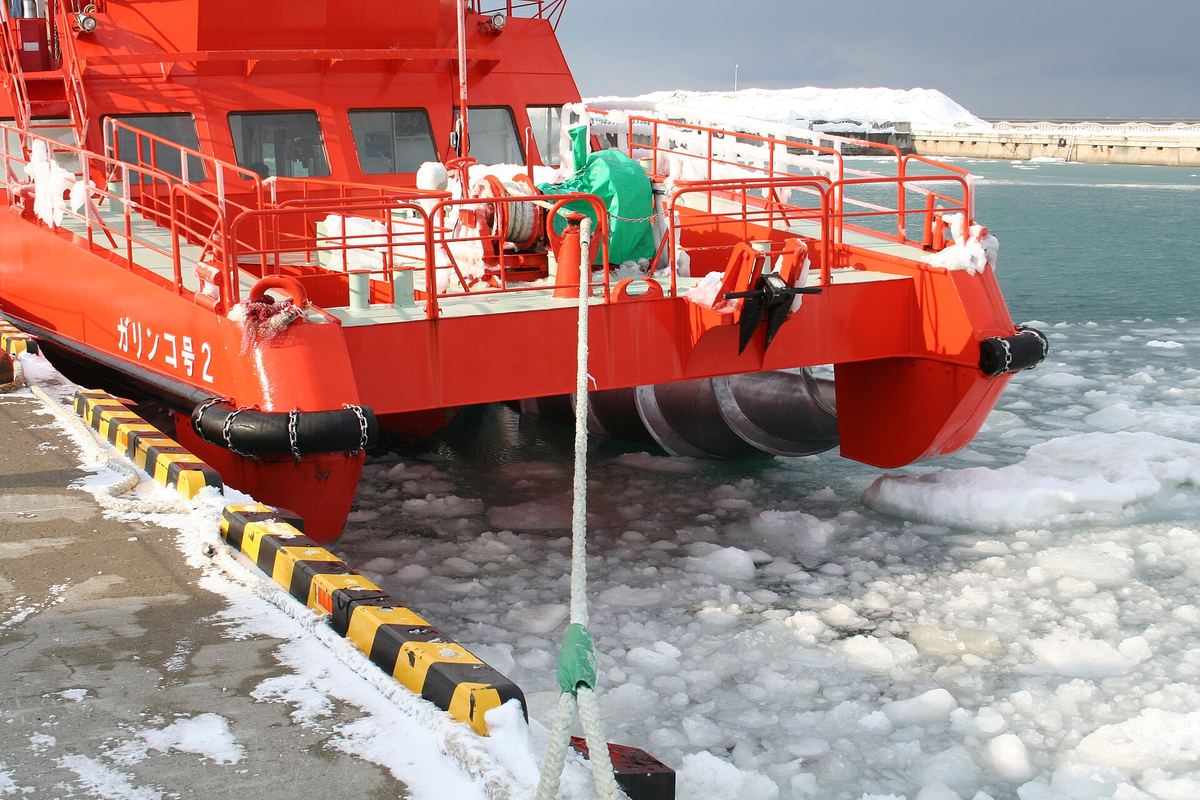 Close-up of the bow of Garinko-go II: a red hull lettered Garinko-go 2 in Japanese, with a silver screw drill under the bow half-submerged in harbor water dotted with broken ice, the ship tied to the quay