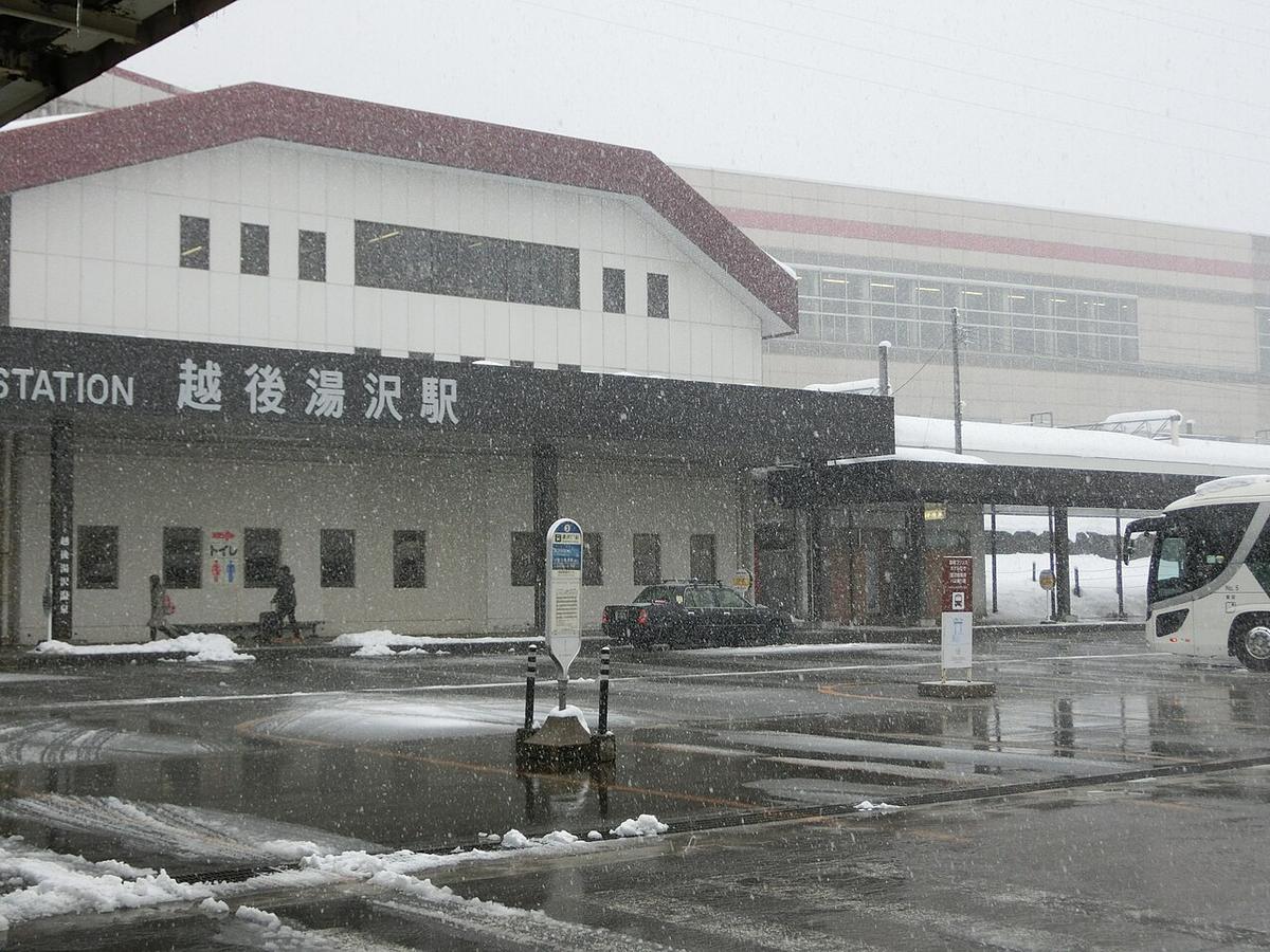 下雪中的越後湯沢站東口:紅色斜屋頂的白色低矮站房,黑色招牌寫著「越後湯沢駅」,後方是新幹線的高架站體;站前廣場濕滑、路邊有積雪,中間立著一根公車站牌,右側停著一輛白色大型巴士,左邊有兩個走過的小小人影