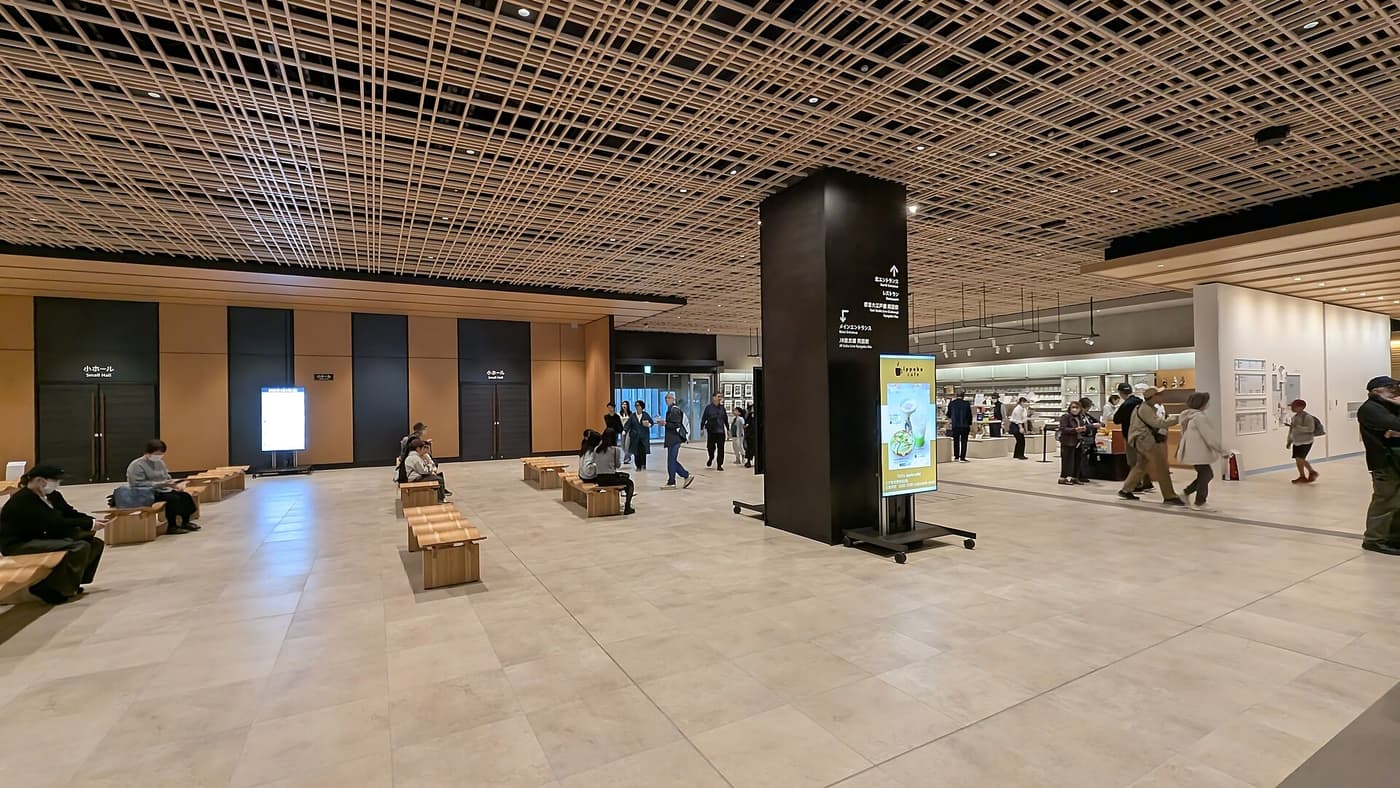 Edo-Tokyo Museum first-floor lobby: a wide pale stone floor under a timber-lattice ceiling, scattered light-wood benches with people sitting on them, a dark square column in the centre carrying arrows to the North Entrance, the restaurant, Toei Oedo Line Ryogoku Station and JR Sobu Line Ryogoku Station, and a bright shop area on the right