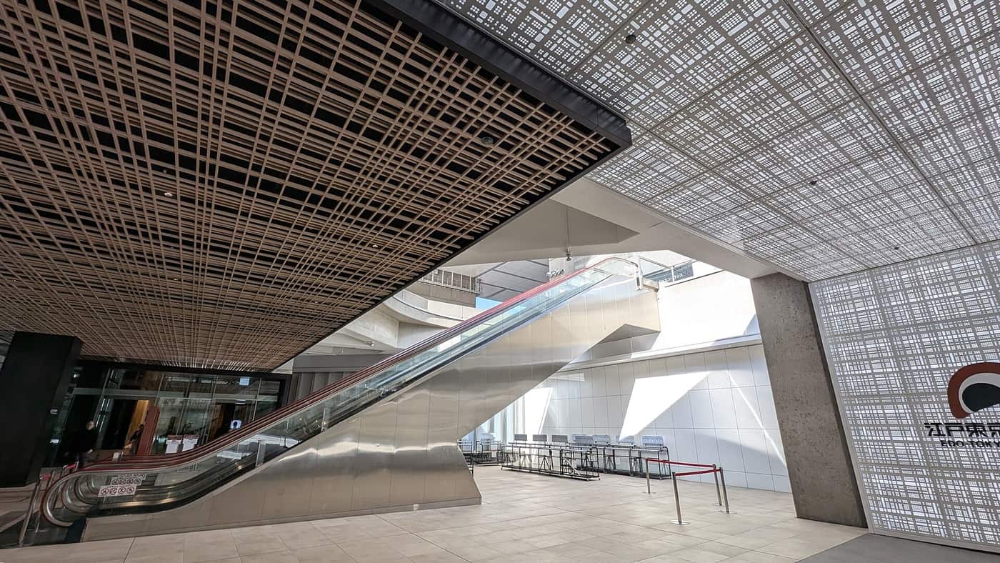Edo-Tokyo Museum first floor: a long silver escalator climbing diagonally to the upper level, a dark timber-lattice ceiling on the left, a white perforated-metal wall carrying the museum's black logo and name on the right, and red rope stanchions set out on the floor below the escalator