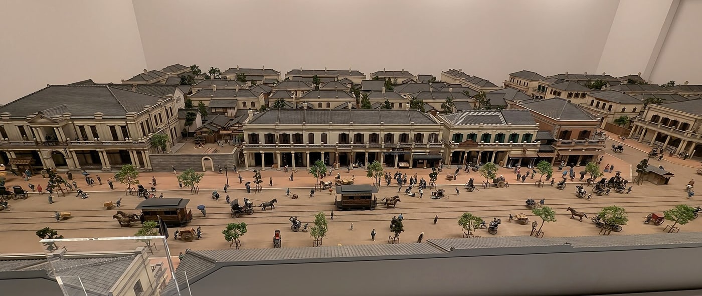 Large-scale model of the Ginza Brick Town inside the Edo-Tokyo Museum permanent exhibition, seen from above: rows of two-storey Western-style brick shophouses lining a very wide earth-coloured street, with horse-drawn tram carriages, rickshaws and several hundred figures in kimono and Western dress