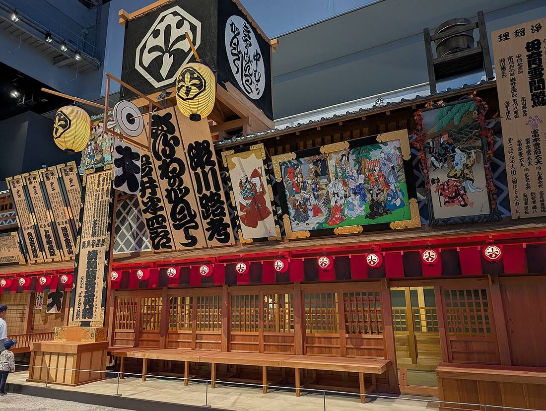 Full-scale reconstruction of the Nakamura-za theatre facade inside the permanent exhibition, with black crest banners above, a row of red lanterns under the eaves, and long wooden benches along the front