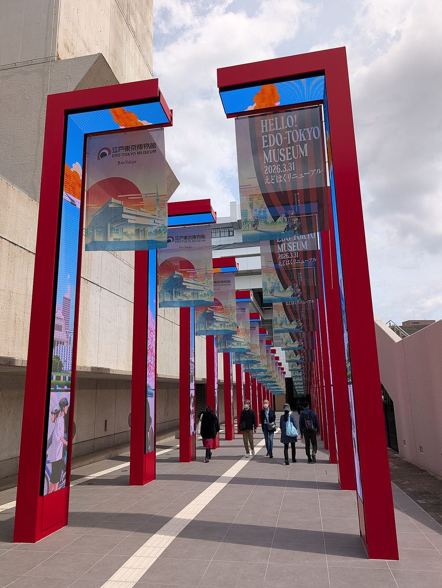 Approach walkway to the Edo-Tokyo Museum entrance: two rows of vermilion gate frames receding into the distance, each holding a vertical screen, with banners reading HELLO! EDO-TOKYO MUSEUM 2026.3.31 hanging between them and five visitors walking down the paving