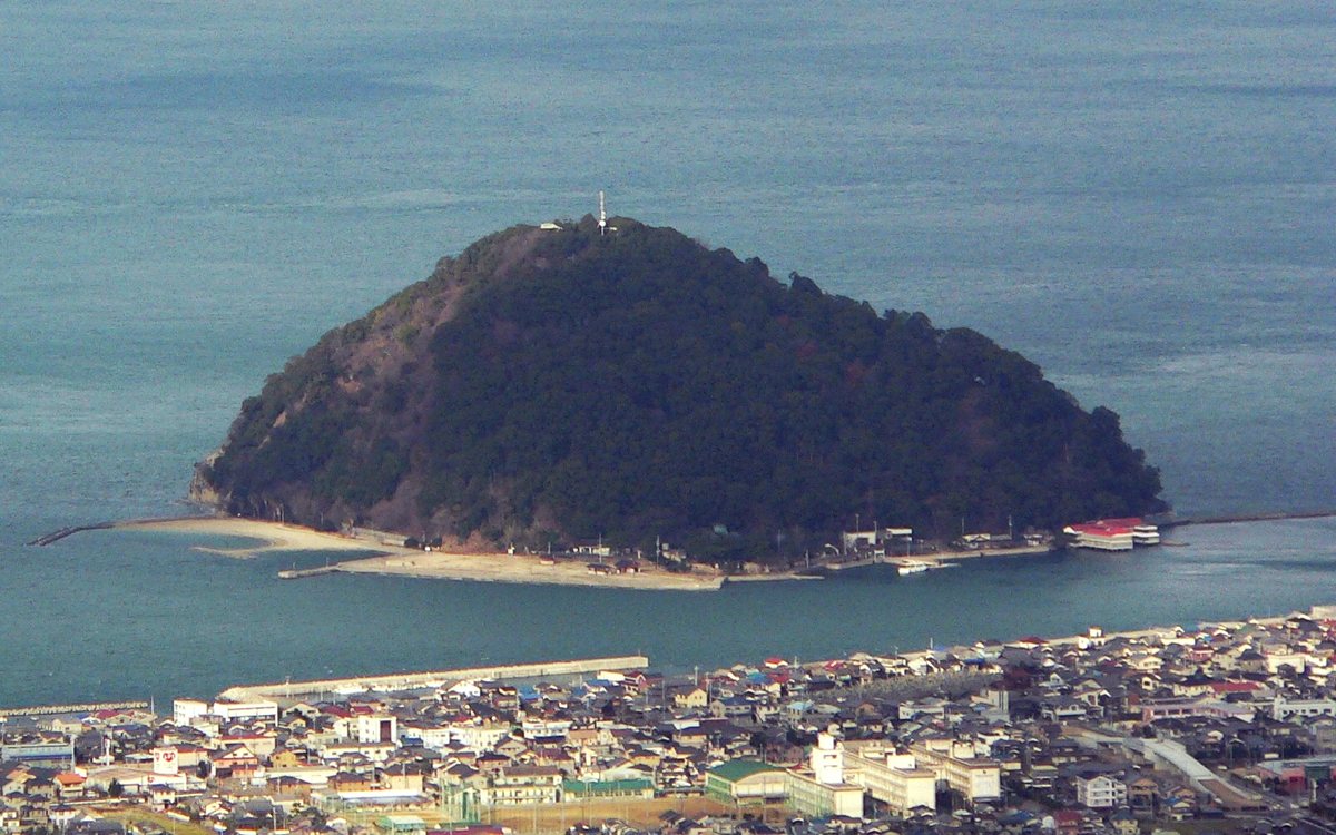 從高處往瀨戶內海看松山市北条:畫面下方是沿海密密麻麻的屋頂與海堤,海面上浮著一座長滿深綠色樹林、圓錐形的小島,島的周圍有一圈白色沙灘