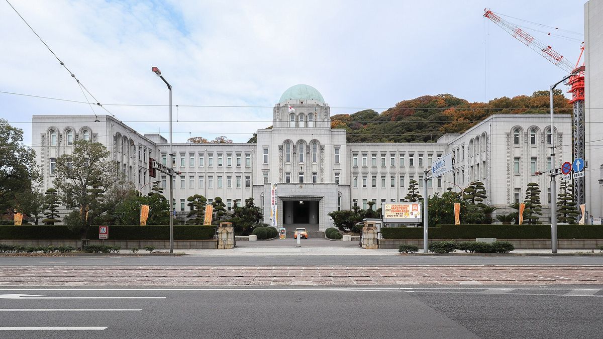 隔著寬闊的馬路看愛媛縣廳本館:左右對稱的白色長條建築,中央塔樓頂著綠色圓頂,正門前有修剪過的松樹,背後是長滿樹的山,馬路上方有電車架線