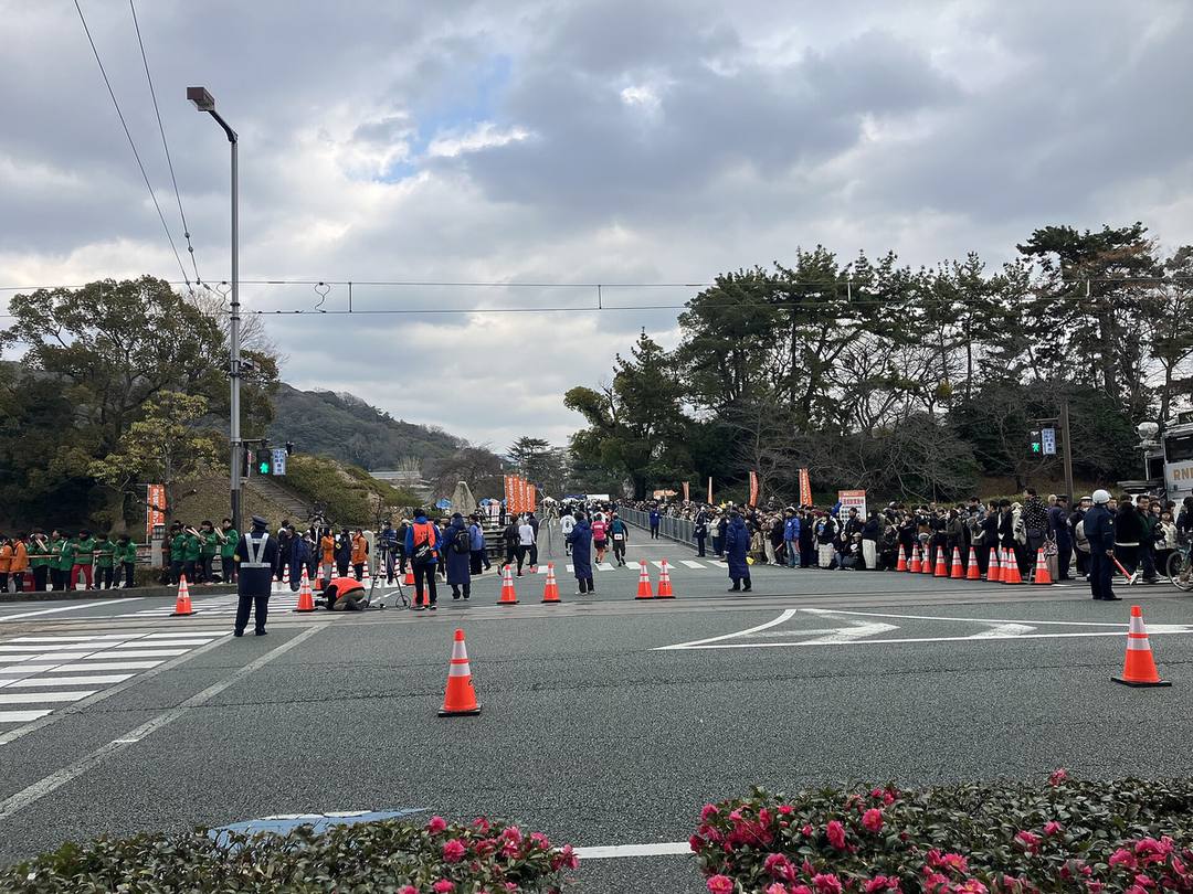 愛媛馬拉松比賽當天的路口:路面有電車軌道、上方拉著電車架線,馬路用橘色三角錐封起來,交通指揮員站在路口,幾位跑者往前方樹林間的會場跑去,兩側人行道擠滿觀眾,左邊是穿綠色外套的加油隊,遠處有寫著「交通規制実施中」的告示牌