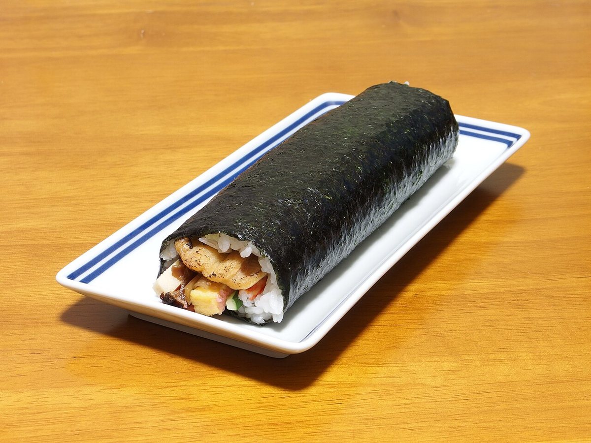 A whole uncut ehomaki futomaki roll on a rectangular white plate, the cut end showing conger eel, rolled omelette and cucumber filling