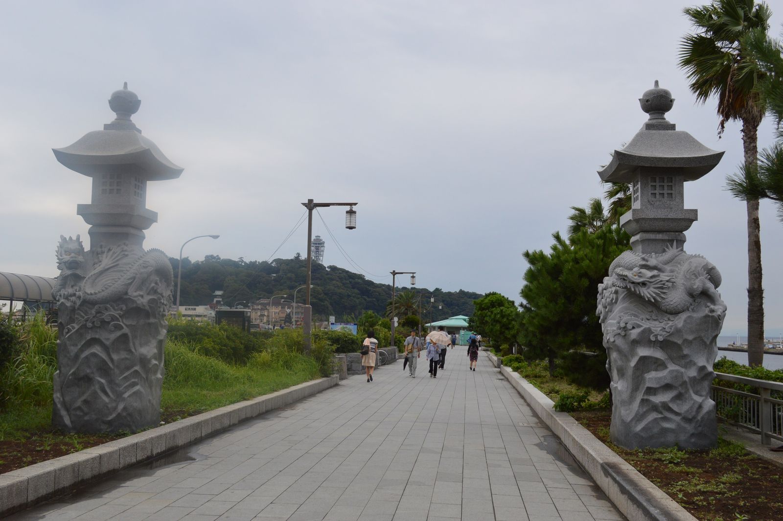 陰天傍晚的江の島弁天橋步道入口:兩側各立一座雕著龍的石燈籠,中間是筆直的石板步道往島上延伸,遠方山頂上看得到 Sea Candle 燈塔,步道上有幾位行人