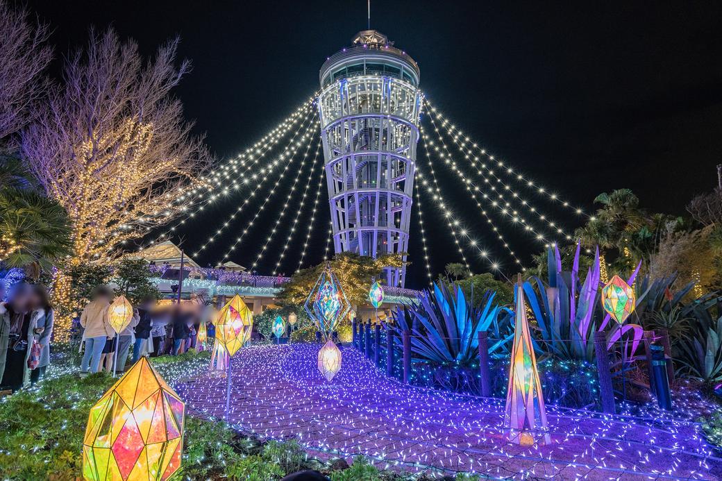 全黑夜空下的 Sea Candle 燈塔,塔身打白光,從塔頂往左右兩側斜拉下數十條白色燈串,像一頂張開的燈傘;前景是藍紫色燈網鋪成的步道與多面體造型的彩色立燈,左側一排遊客在拍照