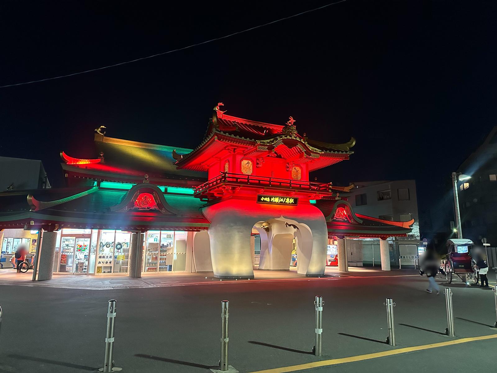 夜裡的小田急片瀬江ノ島站,龍宮城造型的站房屋頂被打上紅色與綠色的燈光,正中央是白色的拱形入口,左邊是亮著燈的便利商店,站前廣場立著一排金屬護欄柱