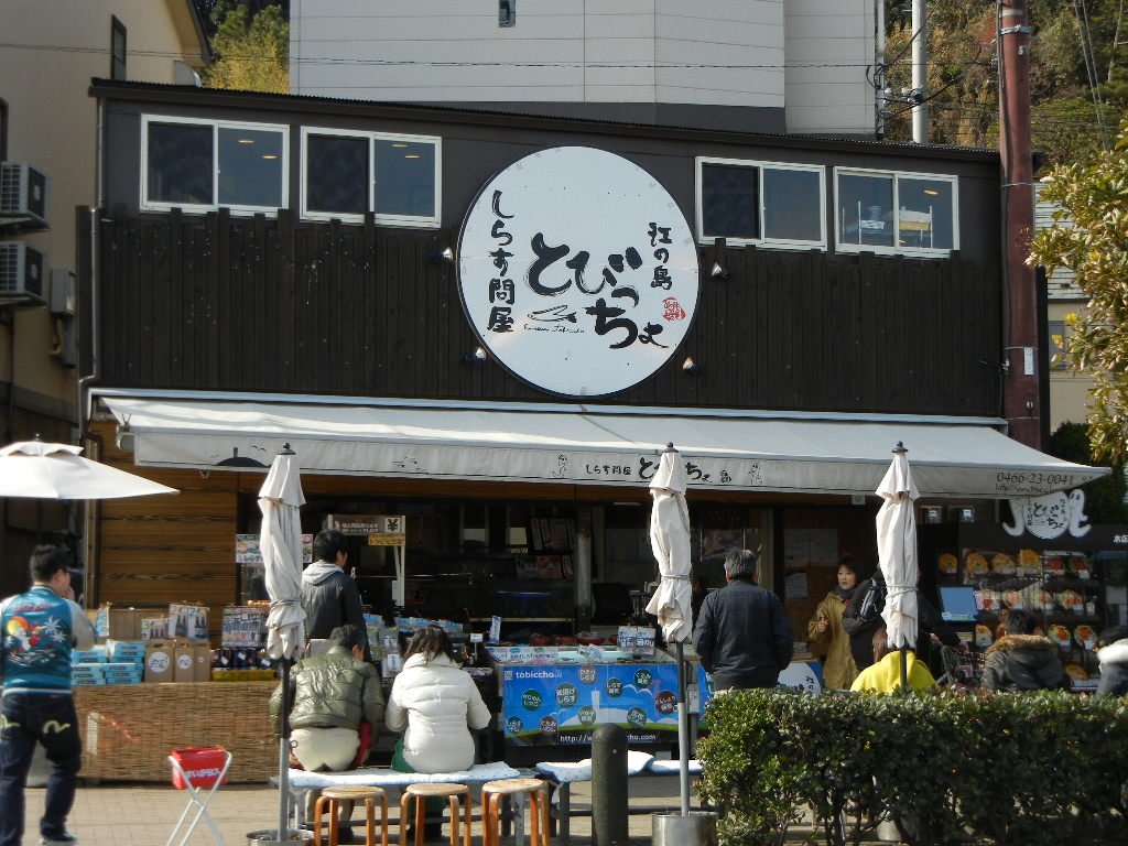 江之島とびっちょ本店的店面，黑色木造外牆掛著白色圓形「しらす問屋 とびっちょ」招牌