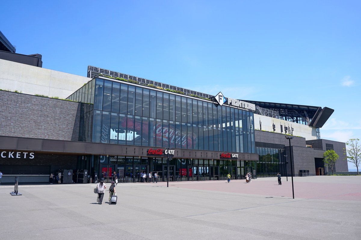 ES CON FIELD HOKKAIDO 的 Coca-Cola GATE:玻璃帷幕入口上方掛著紅色 Coca-Cola GATE 字樣,左邊屋簷下寫著 TICKETS 的是售票處,前方寬廣的廣場上有人拖著行李箱走向入口,晴天