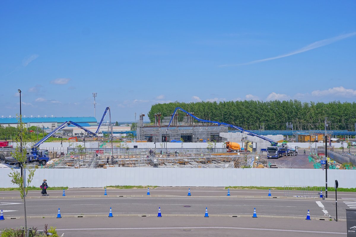 Fビレッジ駅建設工地:白色圍籬後方是鋼筋與模板,兩台藍色長臂混凝土泵車伸在半空,中間是正在灌漿的站體結構,前景是排著藍色三角錐的道路,晴天