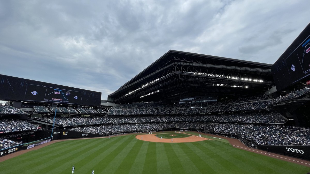 The interior of Es Con Field HOKKAIDO, showing the natural grass field beneath a large open roof structure, tiered seating around the bowl and a glass curtain wall with commercial space beyond the outfield
