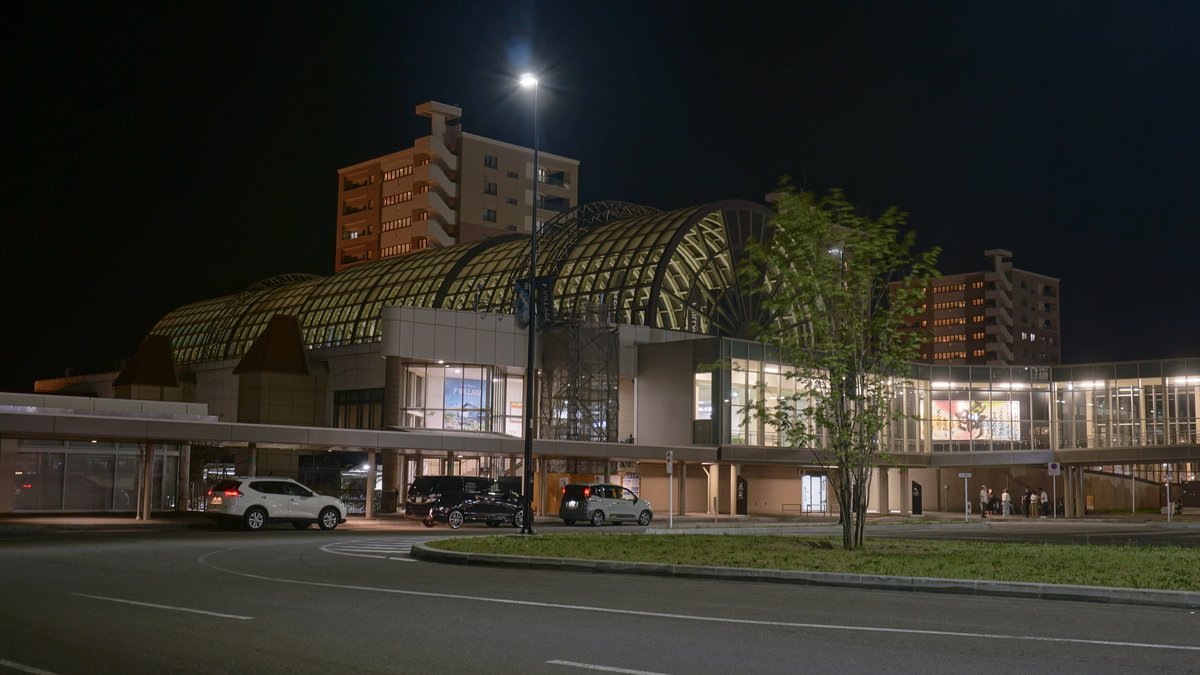 夜間的 JR 北廣島站:亮著燈的拱形玻璃圓頂建築,前面是空曠的站前道路與幾台停著的車,背景有兩棟公寓大樓