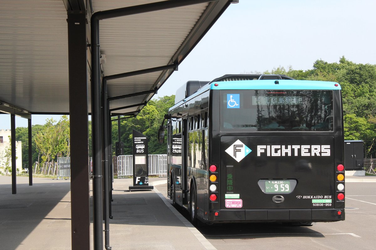 球場南側巴士乘車處:有頂棚的月台旁停著一台黑色車身、藍色車頂的北海道巴士,車尾印著 FIGHTERS 字樣,後窗上方的方向幕顯示北広島駅,月台上立著 BUS 字樣的站牌