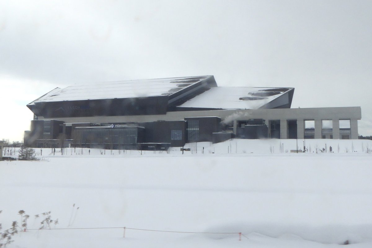 冬天的 ES CON FIELD HOKKAIDO:從 JR 千歲線車窗看過去,前景是一大片積雪的平地,球場的斜面屋頂上也覆著雪,陰天