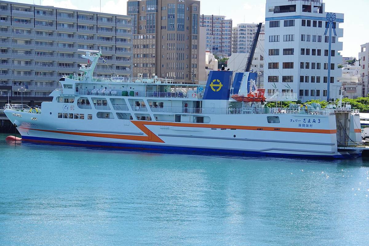 The ferry Zamami 3 berthed at Tomari Port in Naha, a white hull with an orange and dark blue chevron stripe, Zamami Village lettered on the side, Naha apartment blocks behind