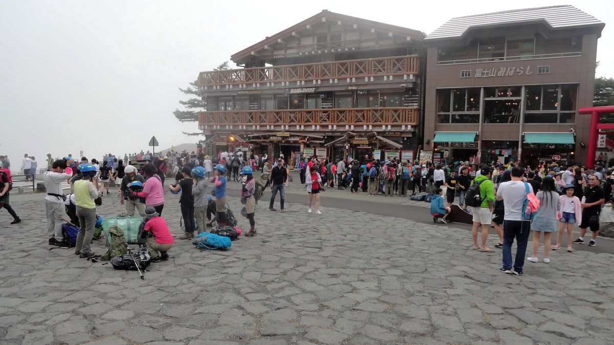 The misty plaza at Fuji Subaru Line 5th Station, crowded in front of timber shops and a restaurant, with helmeted climbers sorting gear beside big packs at left and day trippers in shorts and sandals at right