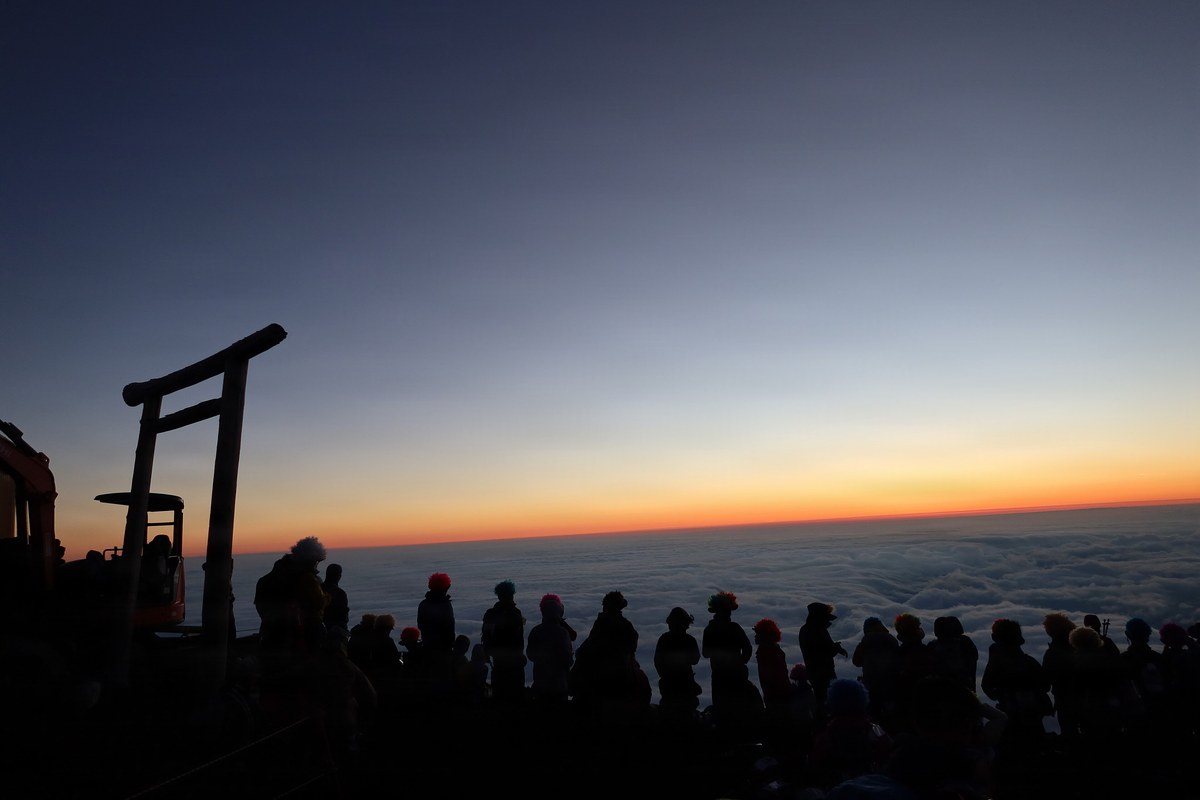 Before sunrise on Mount Fuji's summit, a long line of climbers stands silhouetted along the rim, a wooden torii outlined at left, a sea of clouds stretching to a band of orange light