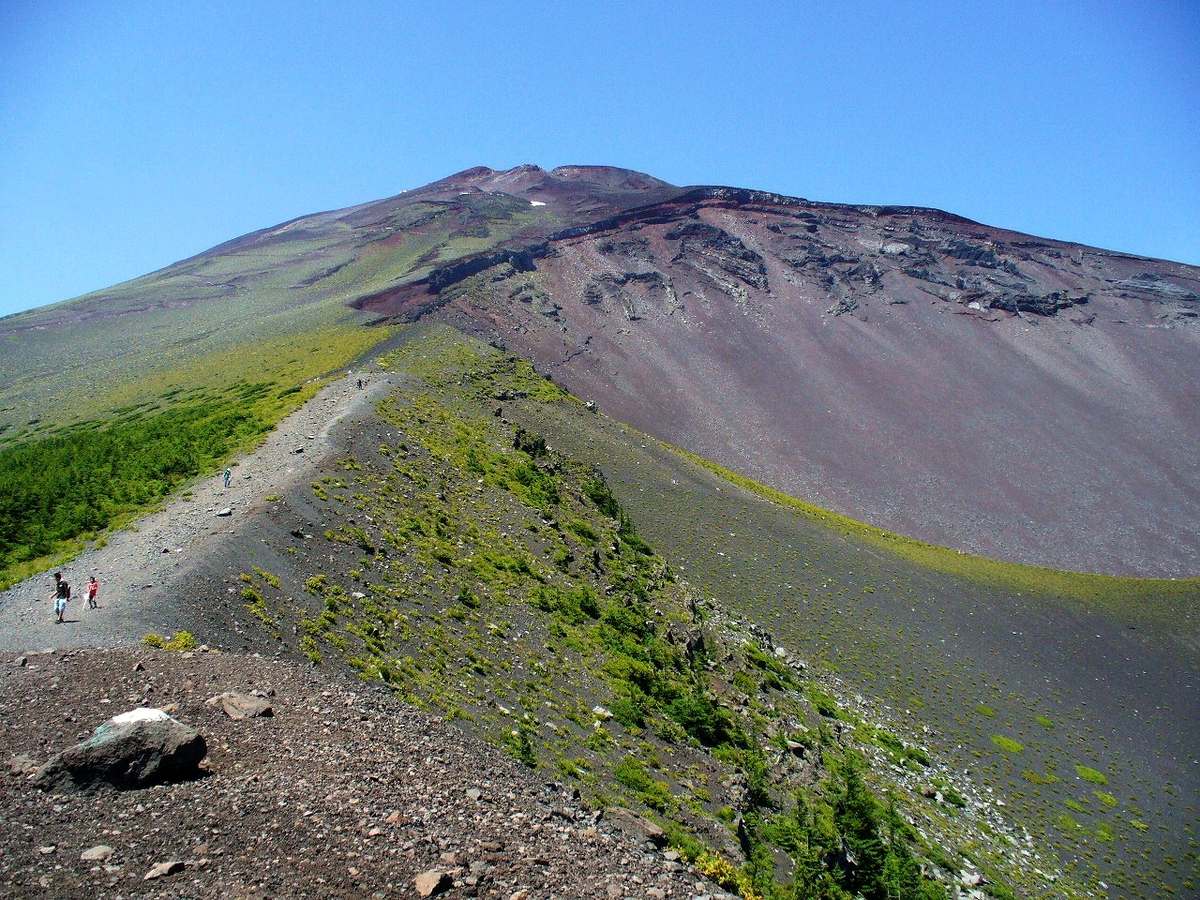 Looking into the Hoei crater from its rim: the right side drops away into a vast red-brown crater wall streaked with dark lava outcrops, while two or three tiny hikers walk the gravel path on the ridge at left