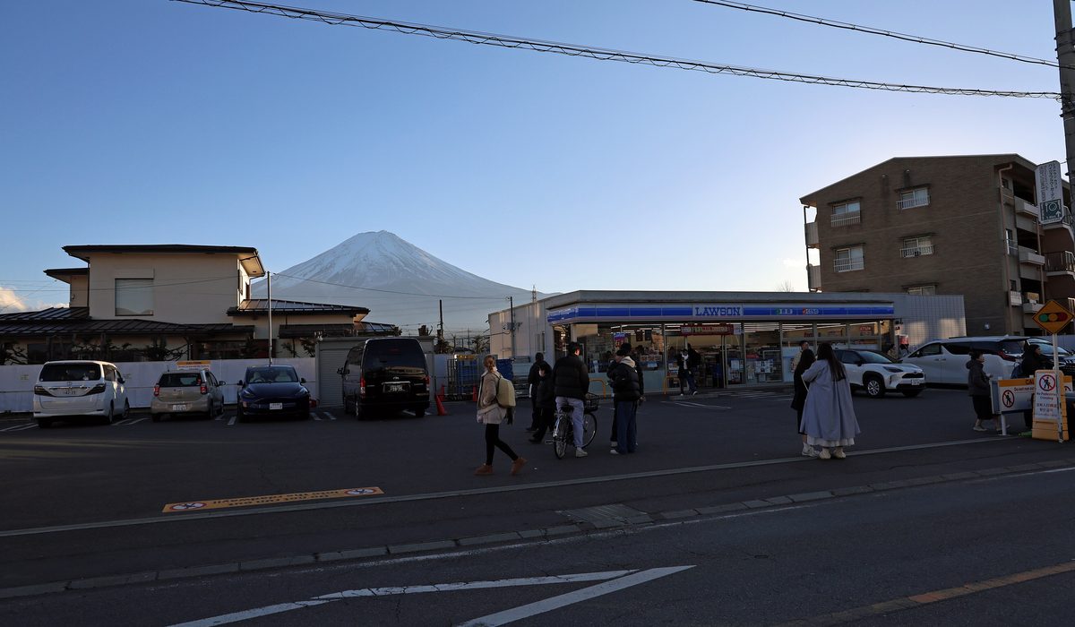 隔著馬路看 LAWSON 河口湖駅前店一帶:左邊是兩層民宅與停車場,富士山從民宅後方露出,右邊是 LAWSON 店面和一棟棕色公寓,店前有幾個行人,右邊路口立著寫有 No Pedestrian Crossing 的告示牌