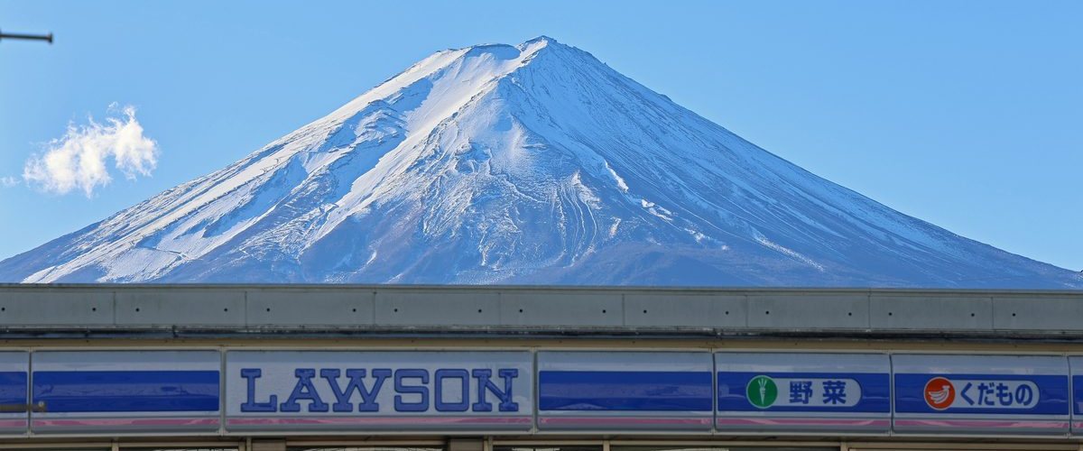 晴天藍天下,山頂積雪的富士山壓在便利商店屋頂正上方,下半是 LAWSON 藍白招牌與「野菜」「くだもの」的店頭橫帶
