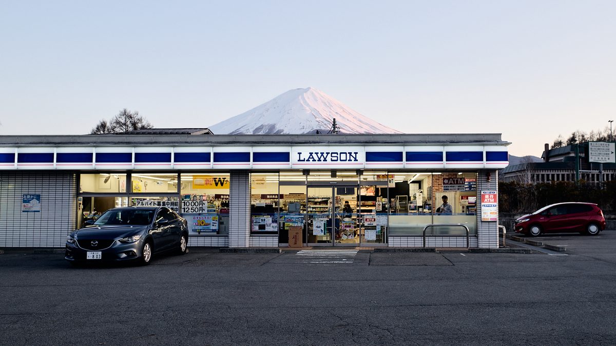 傍晚的另一間 LAWSON:灰白磁磚外牆的店面,屋頂後方是山頂積雪、被夕陽染成淡粉色的富士山,店前空曠的停車場停著兩台車,右邊綠色路標指向富士河口湖町役場
