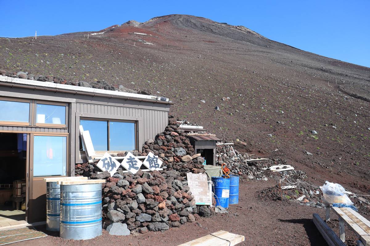 The mountain hut Sunabashirikan on Mount Fuji, its metal-clad building fronted by a wall of stacked black and red volcanic rock, with the bare cinder slope rising behind