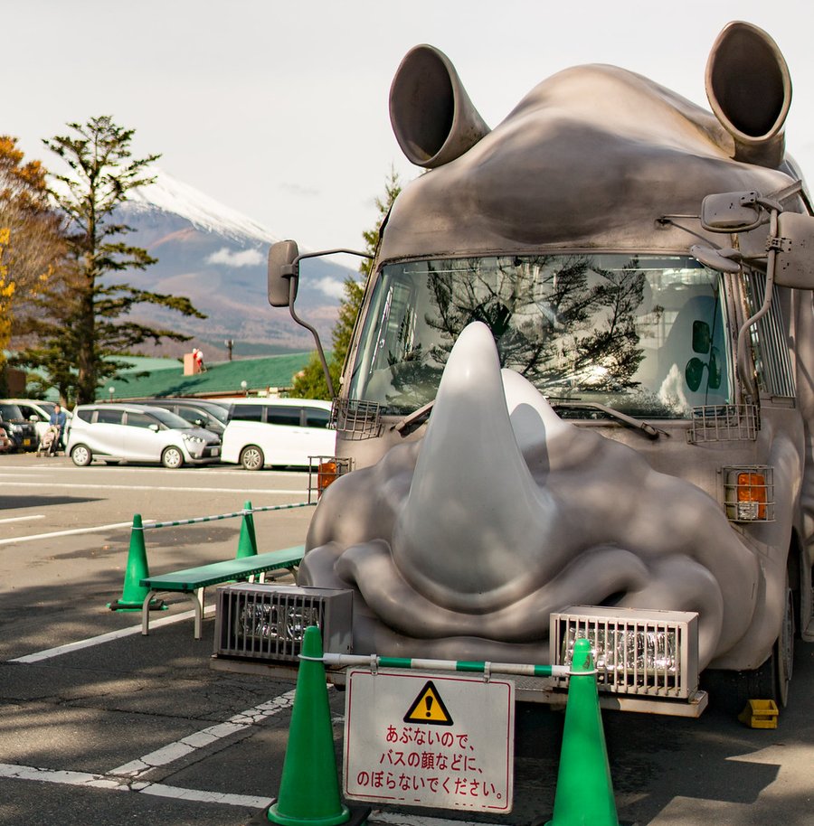 停在富士野生動物園停車場的犀牛造型巴士正面:灰色車頭做成犀牛臉,擋風玻璃下方是一支大犀牛角,車頂兩側有耳朵;車前圍著綠色三角錐與告示牌「あぶないので、バスの顔などに、のぼらないでください」,左後方是積雪的富士山與園區綠色屋頂;2016 年 11 月拍攝