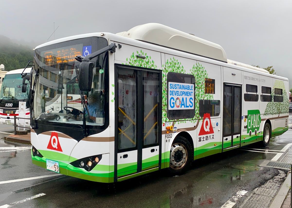 雨天裡停在路邊的富士急巴士 F0053 號車:白色車身、下緣綠色,車側畫著綠色樹木與「SUSTAINABLE DEVELOPMENT GOALS」「EV BUS」字樣,車頭與車側有紅色三角形 Q 字標誌;左後方停著一台京王巴士